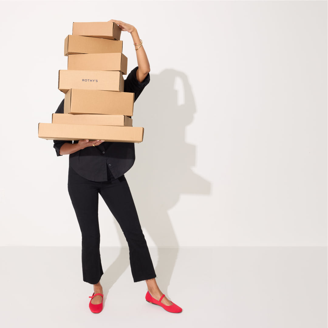 A woman holding a stack of cardboard boxes, wearing The Square Mary Jane in Lollipop Red.