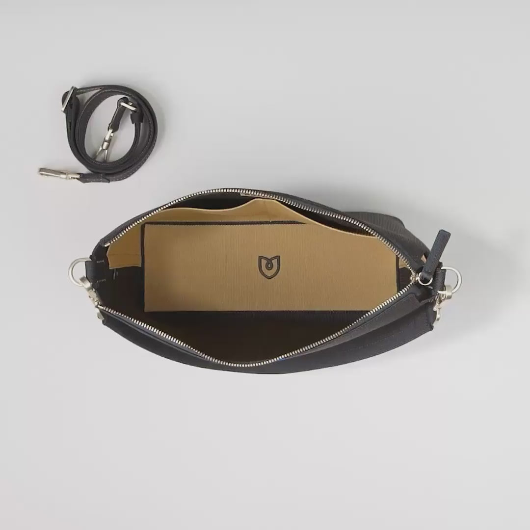 The Daily Crossbody in Ink and Ivory is shown unzipped from the top down, with the adjustable crossbody strap neatly tucked aside. A female model packs the interior slip pockets with sunglasses, lip gloss, hand sanitizer, and Apple AirPods, and then fills the main compartment with a book, an iPhone, and the Mini Universal Pouch. She then zips up the bag, removes the top handle, and hooks the adjustable crossbody strap into place. 