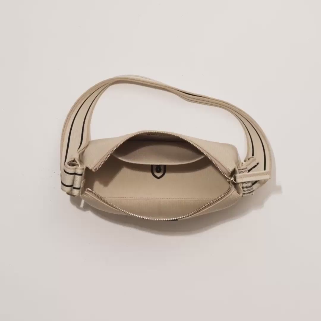 The Casual Crossbody in Parchment shown packed with an iPhone, The Slim Card Case in Grey Houndstooth, makeup, glasses, a notebook, AirPods and lipgloss. A female model then zips up the bag and carries it out of frame.